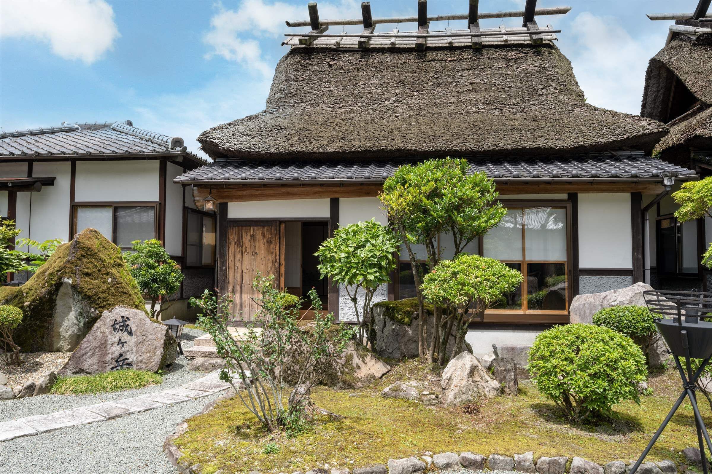 Un edificio japonés tradicional con techo de paja, rodeado de un jardín cuidado con piedras, musgo y diversas plantas.