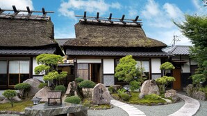 Un edificio tradicional japonés con techo de paja, rodeado de un jardín cuidado con rocas, árboles y un sendero de piedra.
