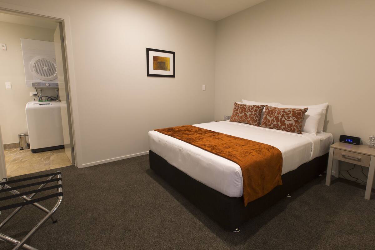 Una habitación de hotel con una cama, una mesita de noche, una pequeña mesa, una silla, un cuadro en la pared y una lavadora en la habitación contigua.