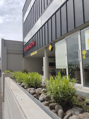 Un bâtiment moderne, orné d’une enseigne « RAMADA », doté d’une entrée en verre et agrémenté d’un espace paysager avec des plantes et des rochers.