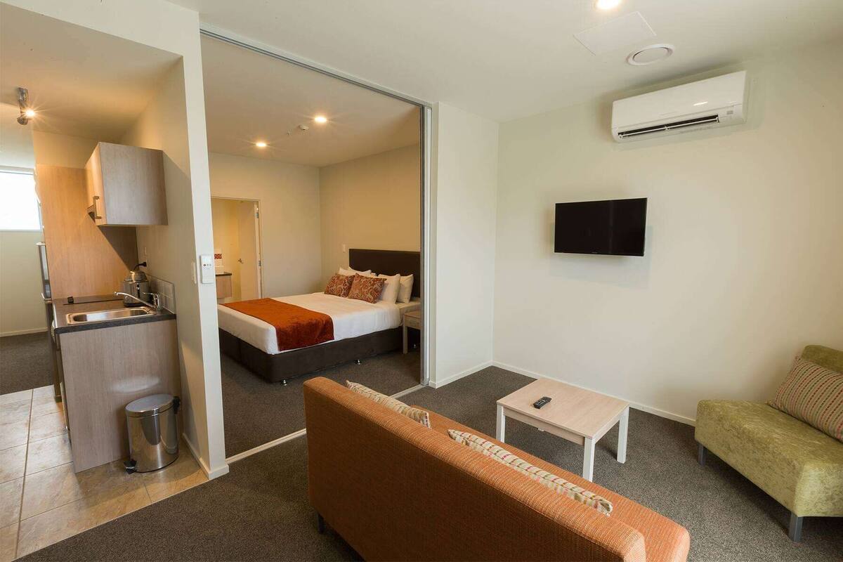 Habitación de hotel con cama, televisor, sofá, una mesita, zona de cocina y aire acondicionado.