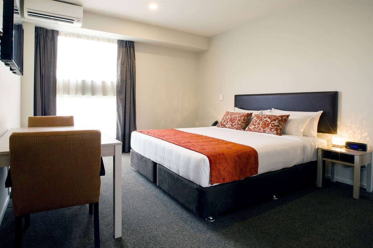 Una habitación de hotel con cama, escritorio, silla, mesita de noche y unidad de aire acondicionado.