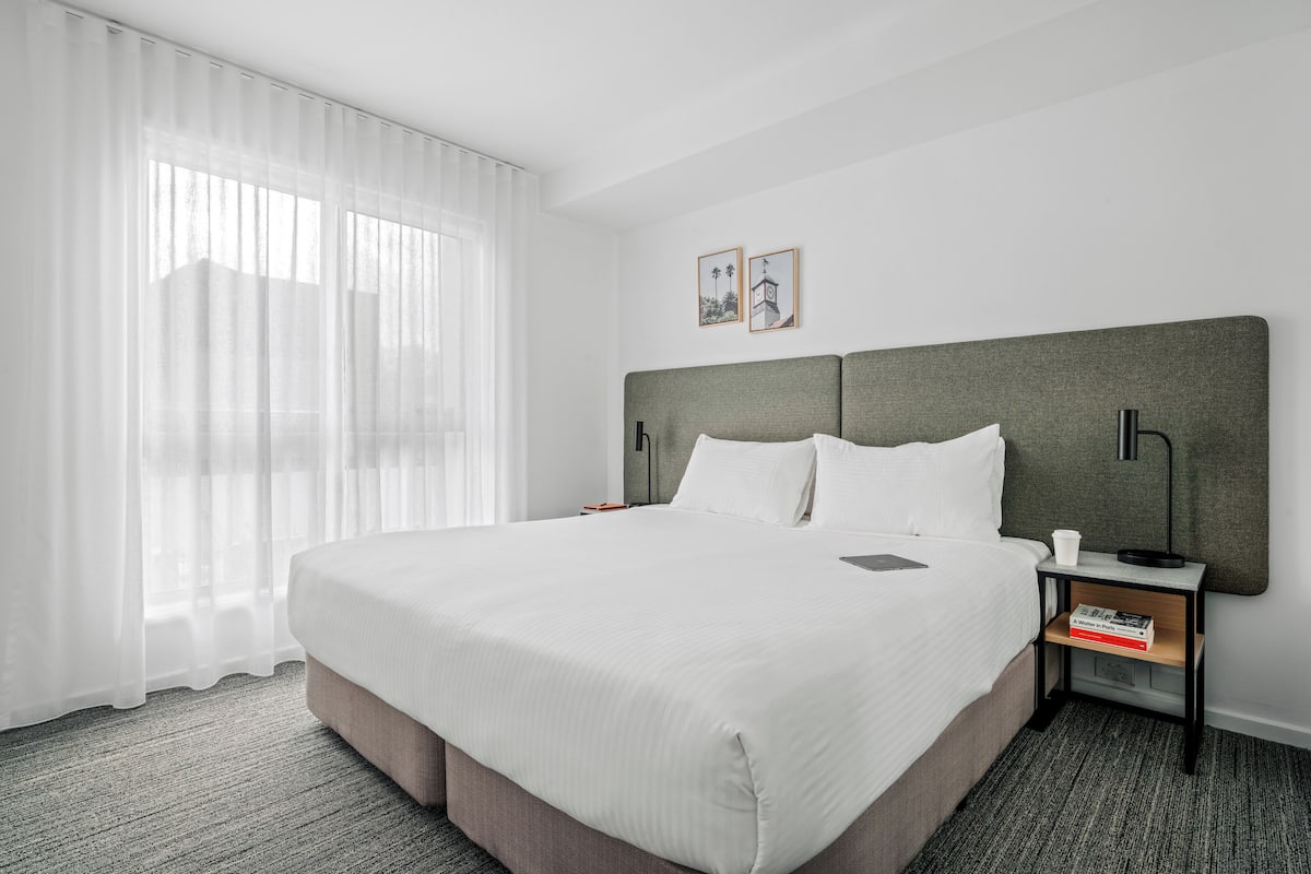 Uma cama bem arrumada com lençóis brancos, cabeceira cinzenta e mesa de cabeceira com abajur e um livro.