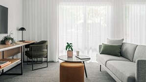 Uma sala de estar moderna com sofá, mesa de centro redonda, escrivaninha com cadeira, televisão e uma grande janela com cortinas leves.