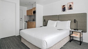 Quarto de hotel moderno com uma cama grande, mesa de cabeceira com luminária, pequena área de cozinha e porta à esquerda.