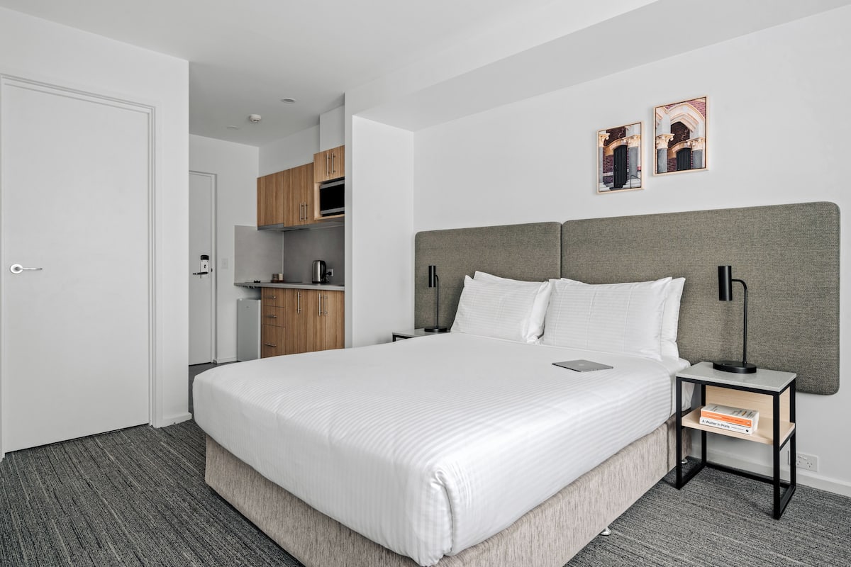 Quarto de hotel moderno com uma cama grande, mesa de cabeceira com luminária, pequena área de cozinha e porta à esquerda.