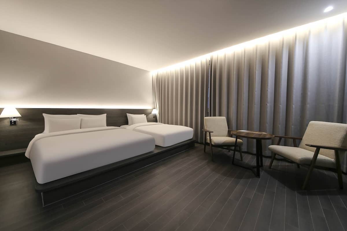 A modern hotel room with a large bed, two bedside tables, a seating area with two chairs and a small table, and a large window with curtains.