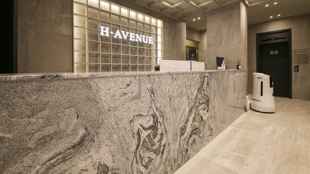 Un área de recepción con mostrador de mármol, un muro de cristal con el letrero 'H. AVENUE', un mostrador de recepción y un dispensador de gel antibacterial.