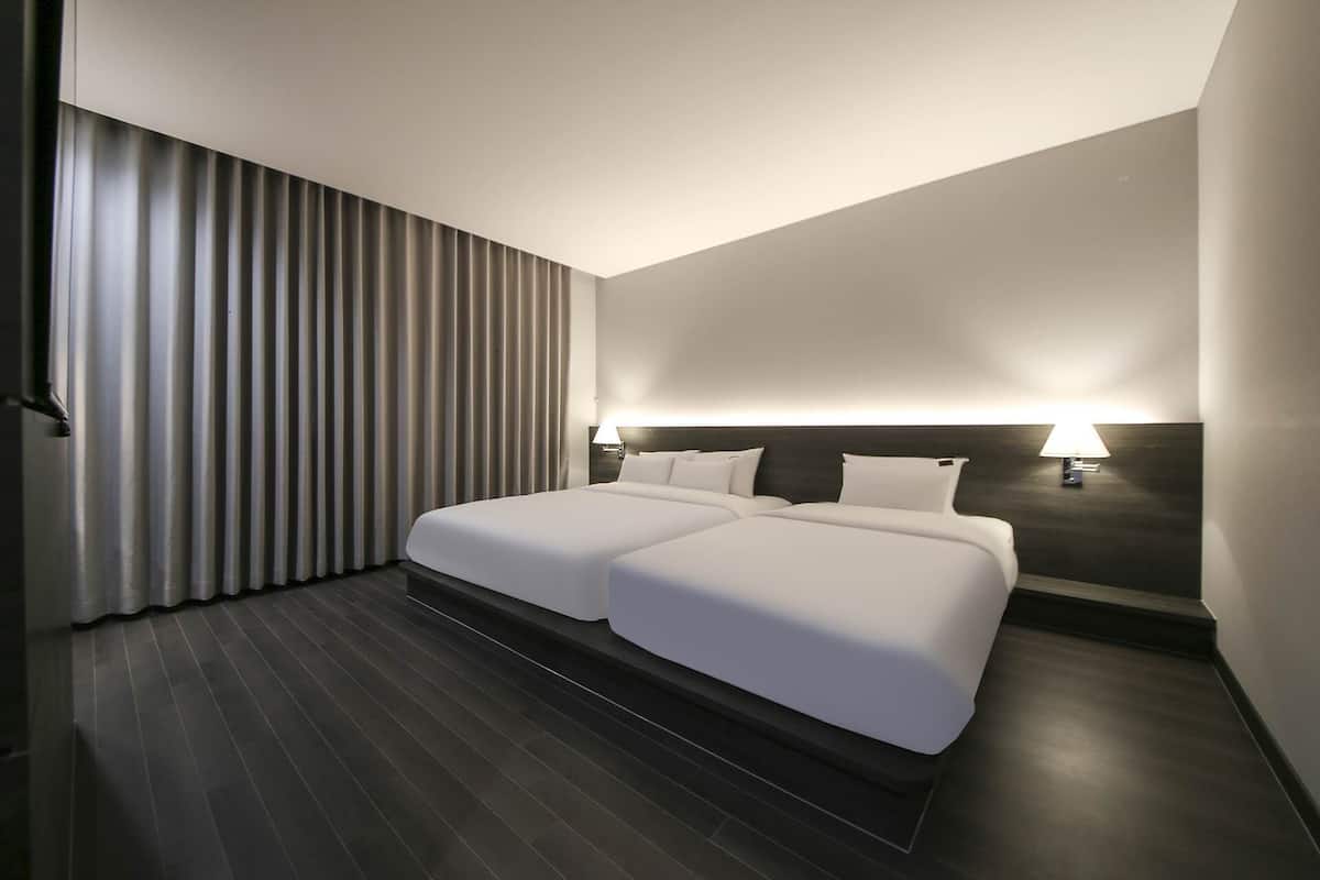 A modern hotel room with two beds, dark wooden flooring, and a large window with vertical blinds.