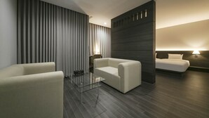 Una habitación de hotel moderna con piso de madera oscura, una cama grande con cabecera, dos sillones blancos, una mesa de centro de vidrio y un ventanal con persianas verticales.
