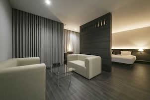 Una habitación de hotel moderna con piso de madera oscura, una cama grande con cabecera, dos sillones blancos, una mesa de centro de vidrio y un ventanal con persianas verticales.