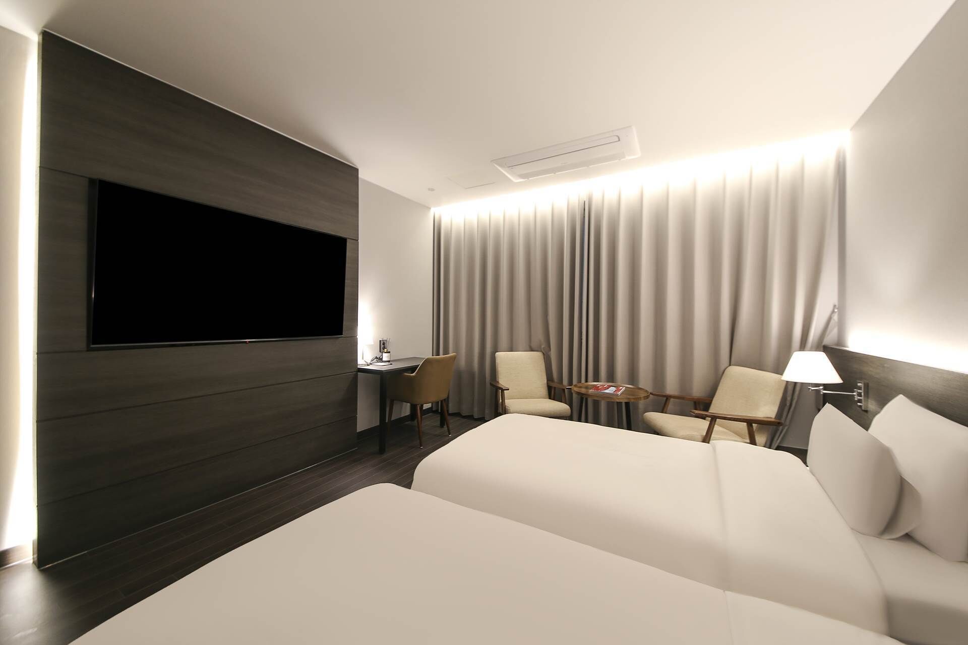 A modern hotel room with a large bed, a desk, two chairs, a TV, and a curtain backdrop.
