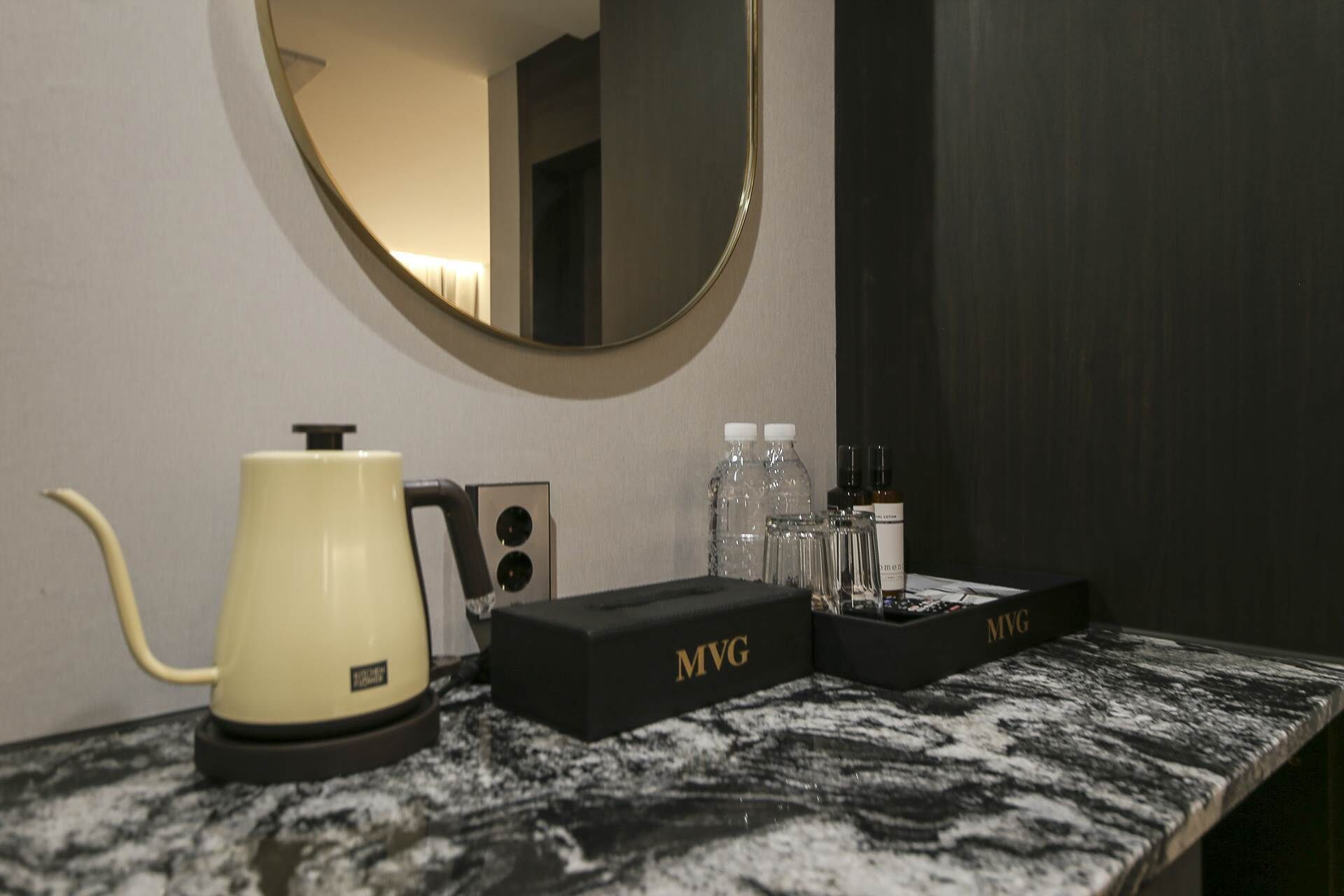 Une salle de bain avec un plan de toilette en marbre, une bouilloire électrique jaune, une boîte noire portant l’inscription MVG et un miroir avec un cadre doré.