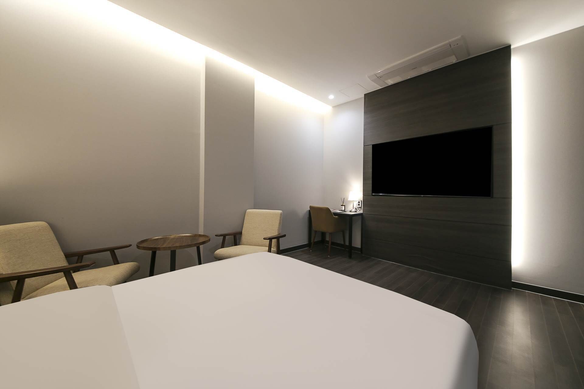 A hotel room with a large bed, two armchairs, a small table, a desk with a lamp, and a mounted TV.
