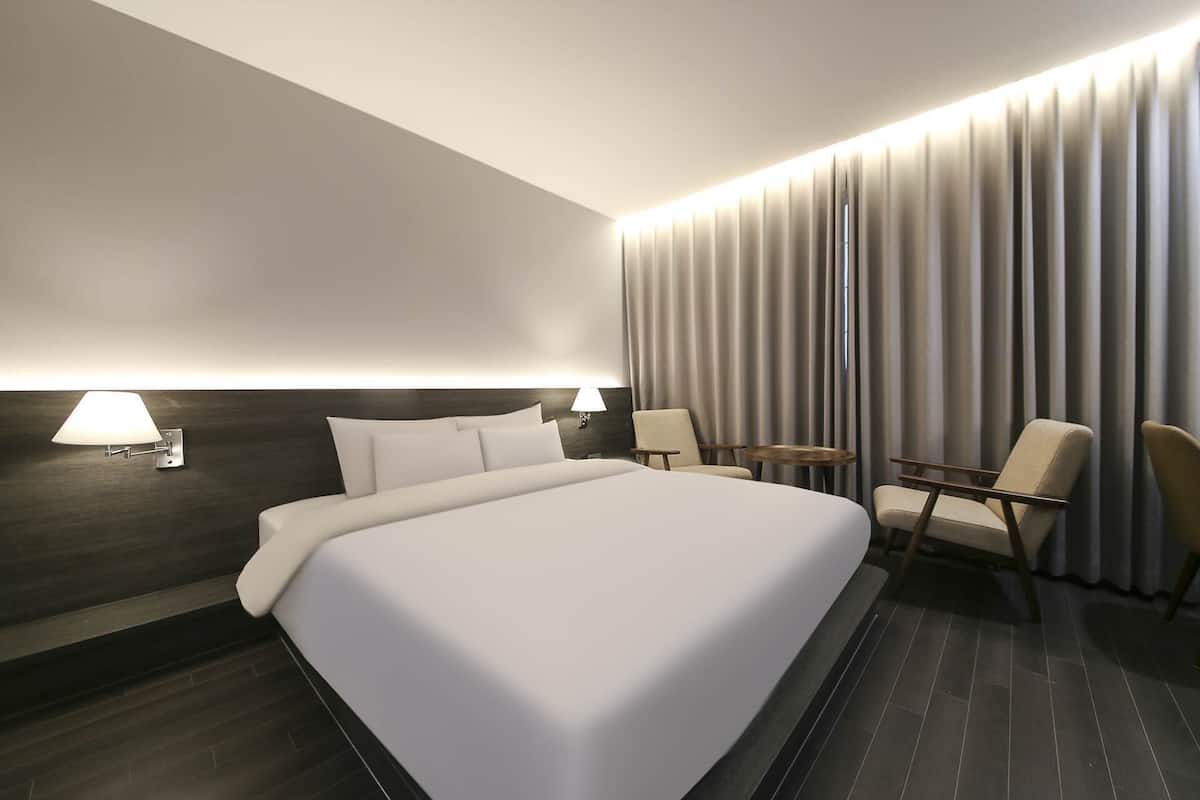 A modern hotel room with a large bed, two armchairs, a small table, and a wall-mounted lamp.