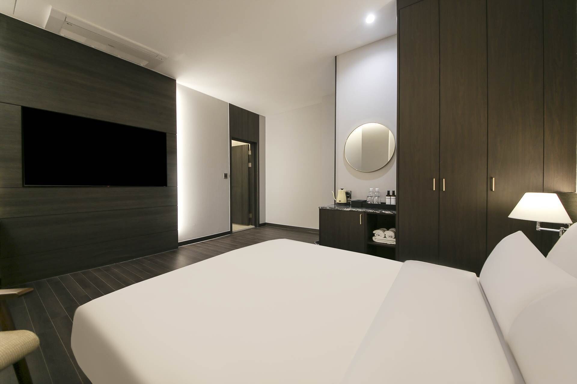 A modern hotel room with a large bed, a flat-screen TV mounted on the wall, a round mirror above the dresser, and a nightstand with a lamp.