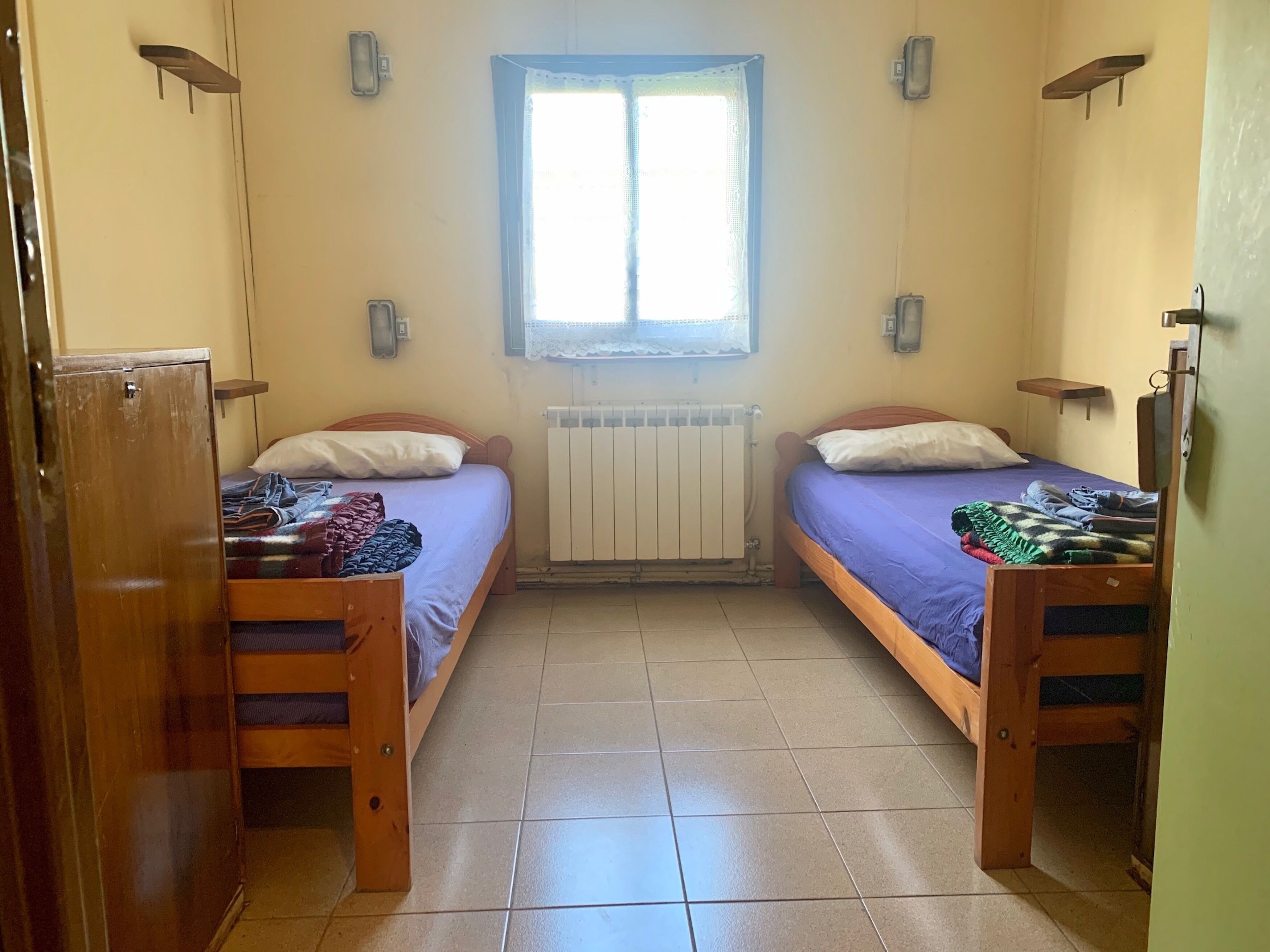 A small, well-lit room with two wooden beds, a window with lace curtains, a radiator, and a wooden wardrobe.