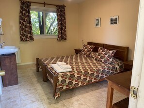 Ein Schlafzimmer mit Bett, Nachttisch, Fenster mit Vorhängen, einem Wandbild und einem Badezimmer mit Waschbecken und Spiegel.