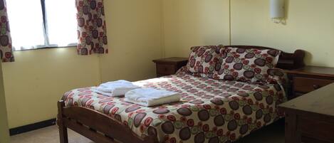 A bedroom with a bed, a nightstand, a bedside table, a window with curtains, and a wall-mounted light.