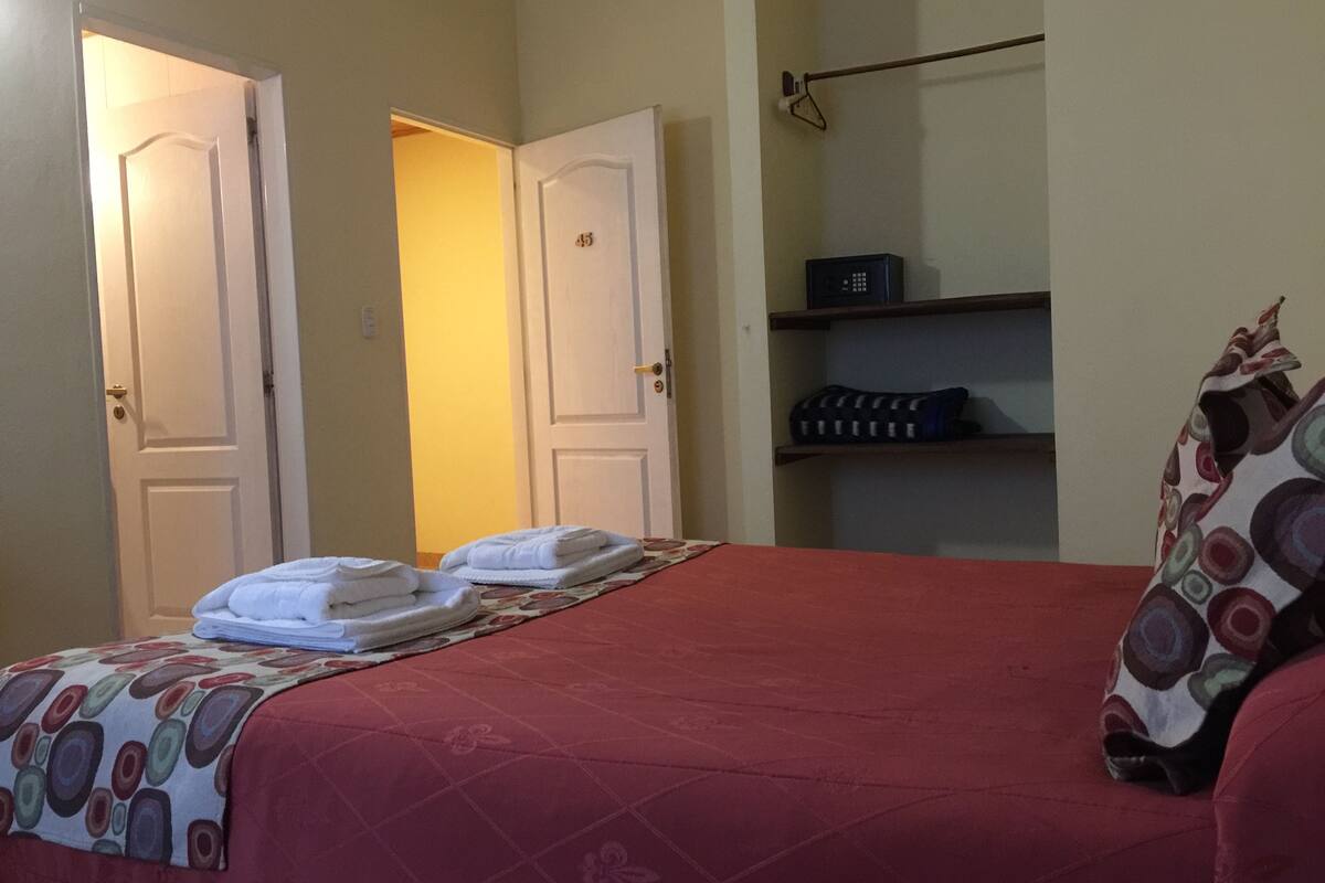Une chambre d’hôtel avec un lit, deux portes, une étagère avec quelques objets et une couvre-lit à motifs.