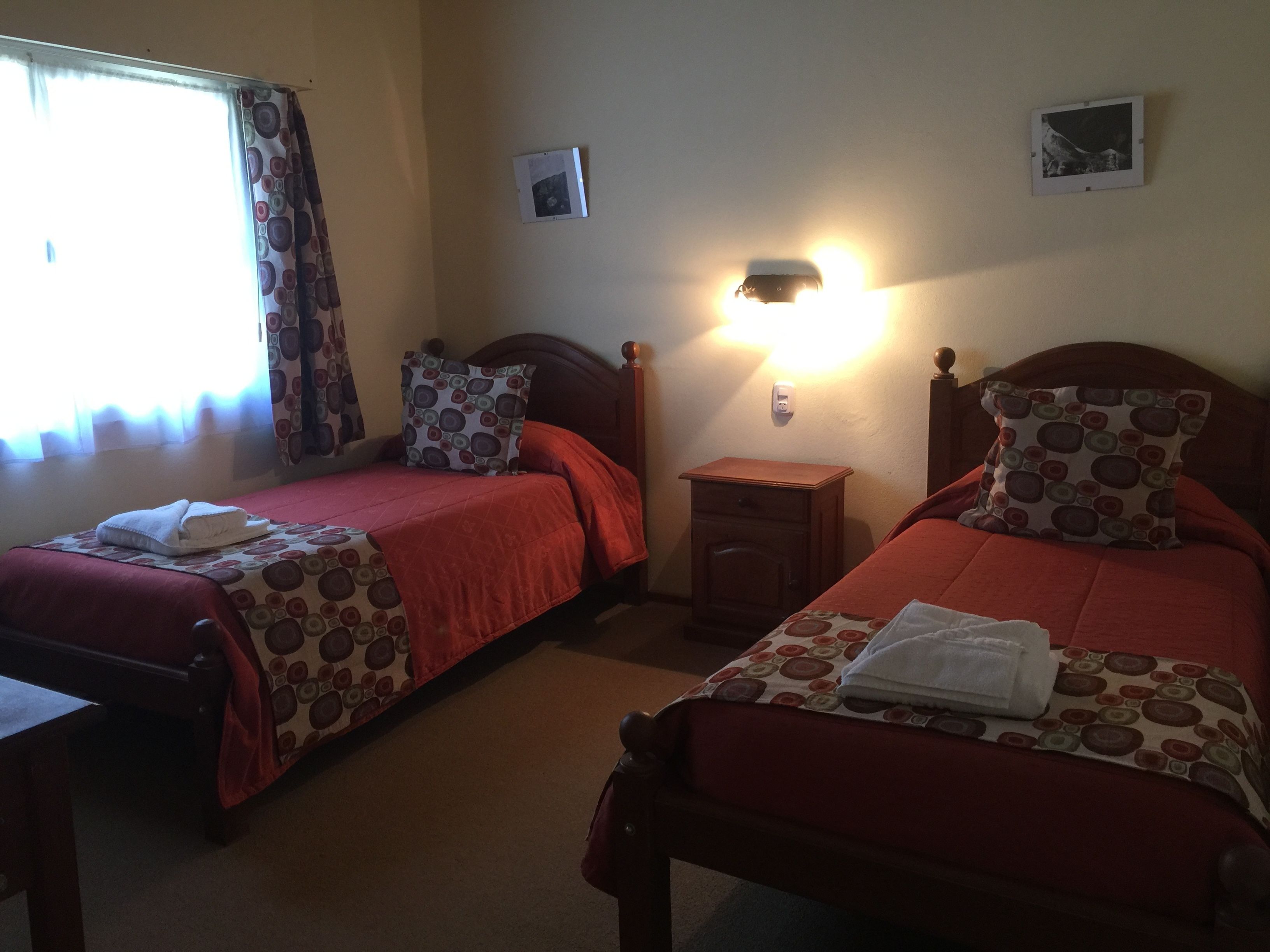 Two single beds with red bedding and matching pillows, a bedside table, a nightstand, a wall-mounted light, and a window with curtains.