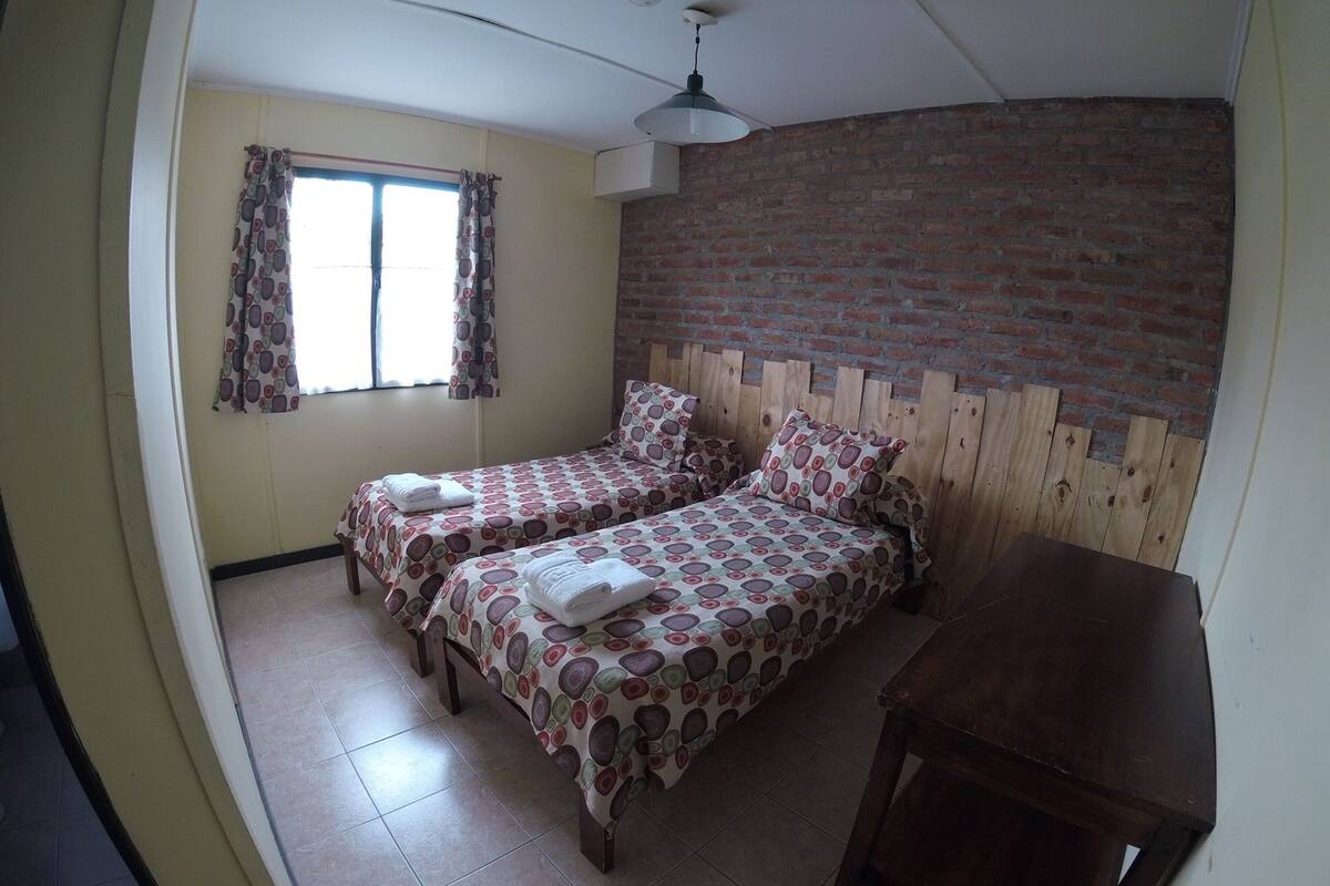 Une chambre avec deux lits, un mur en briques, une fenêtre avec des rideaux et un ventilateur de plafond.