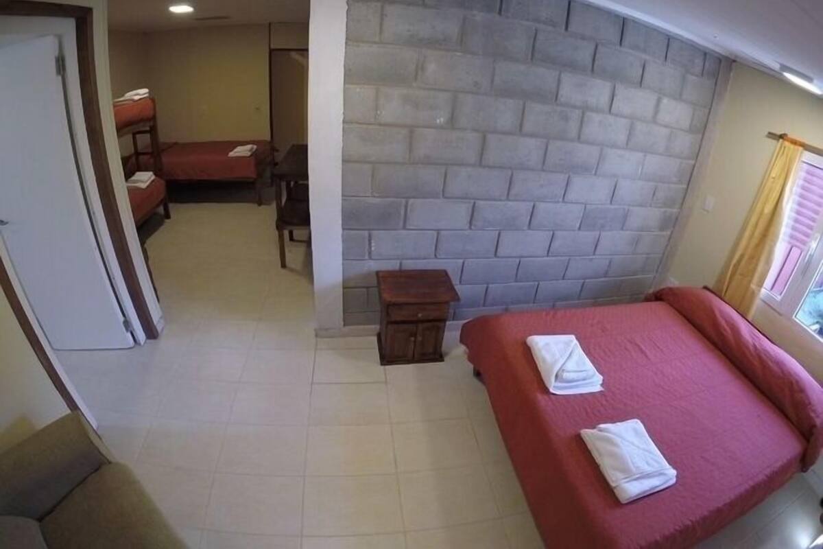 Une chambre avec un mur en briques, un lit avec une literie rouge, une petite table de chevet en bois et une fenêtre avec des rideaux.
