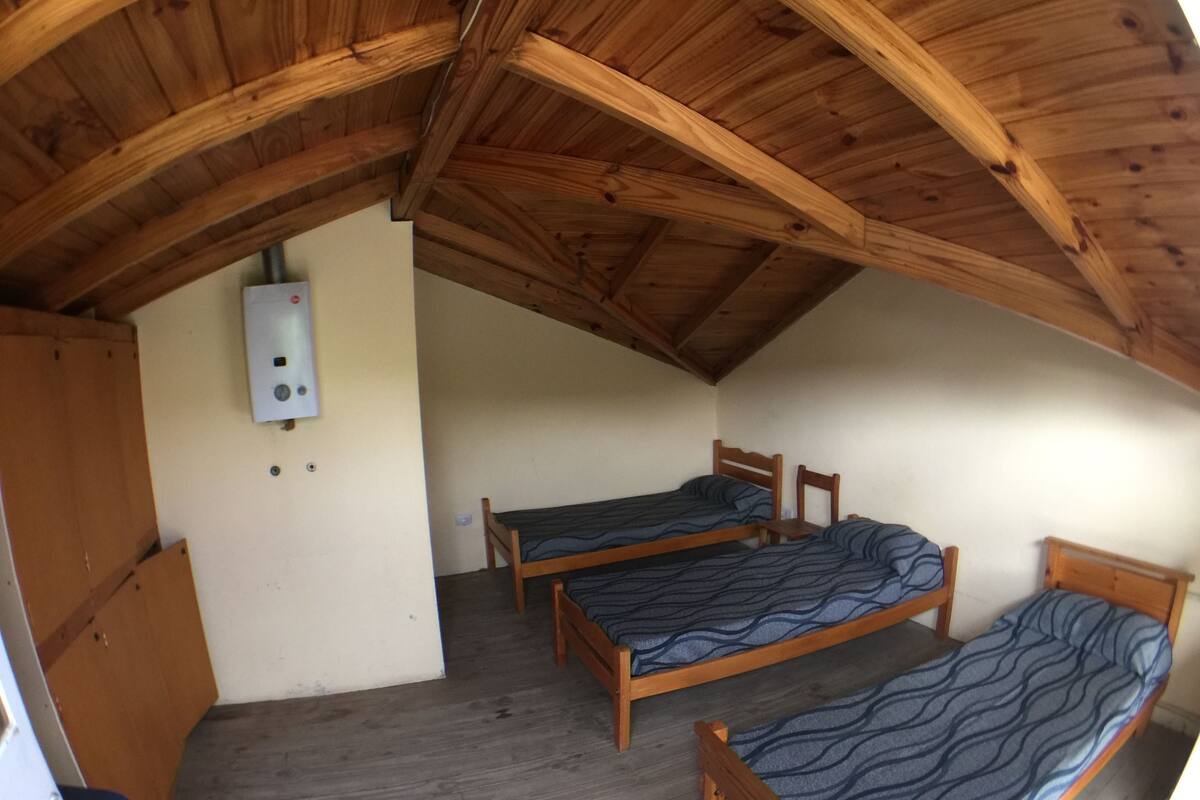 Une petite chambre mansardée avec un toit en bois, comprenant trois lits simples, un radiateur et une porte en bois.