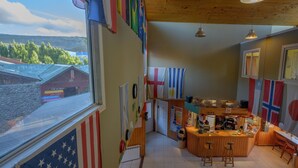 A room with a wooden ceiling, a window with an American flag, a small bar area, and various flags on the walls. - Hostel del Glaciar Pioneros (El Calafate)