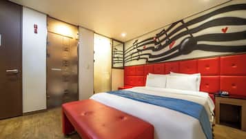 A hotel room with a red upholstered headboard, a large bed with white and blue bedding, a bench at the foot of the bed, and a wall with a musical note design.