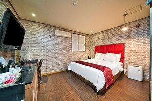 Habitación de hotel con pared de ladrillo, cama con cabecera roja, escritorio con ordenador y ventana con persianas.