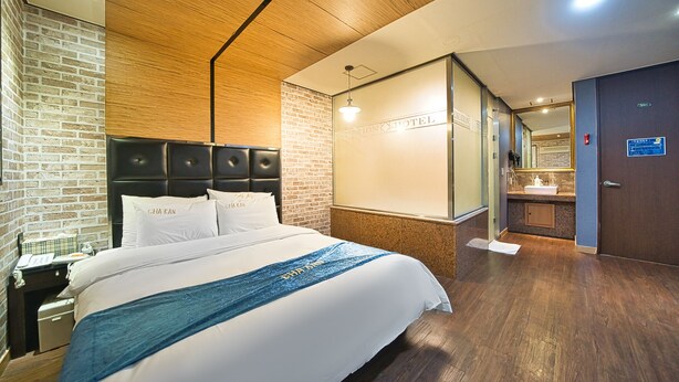 A hotel room with a brick wall, a wooden ceiling, a bed with white linens, a blue decorative pillow, a nightstand with a telephone, and a bathroom with a sink and mirror.