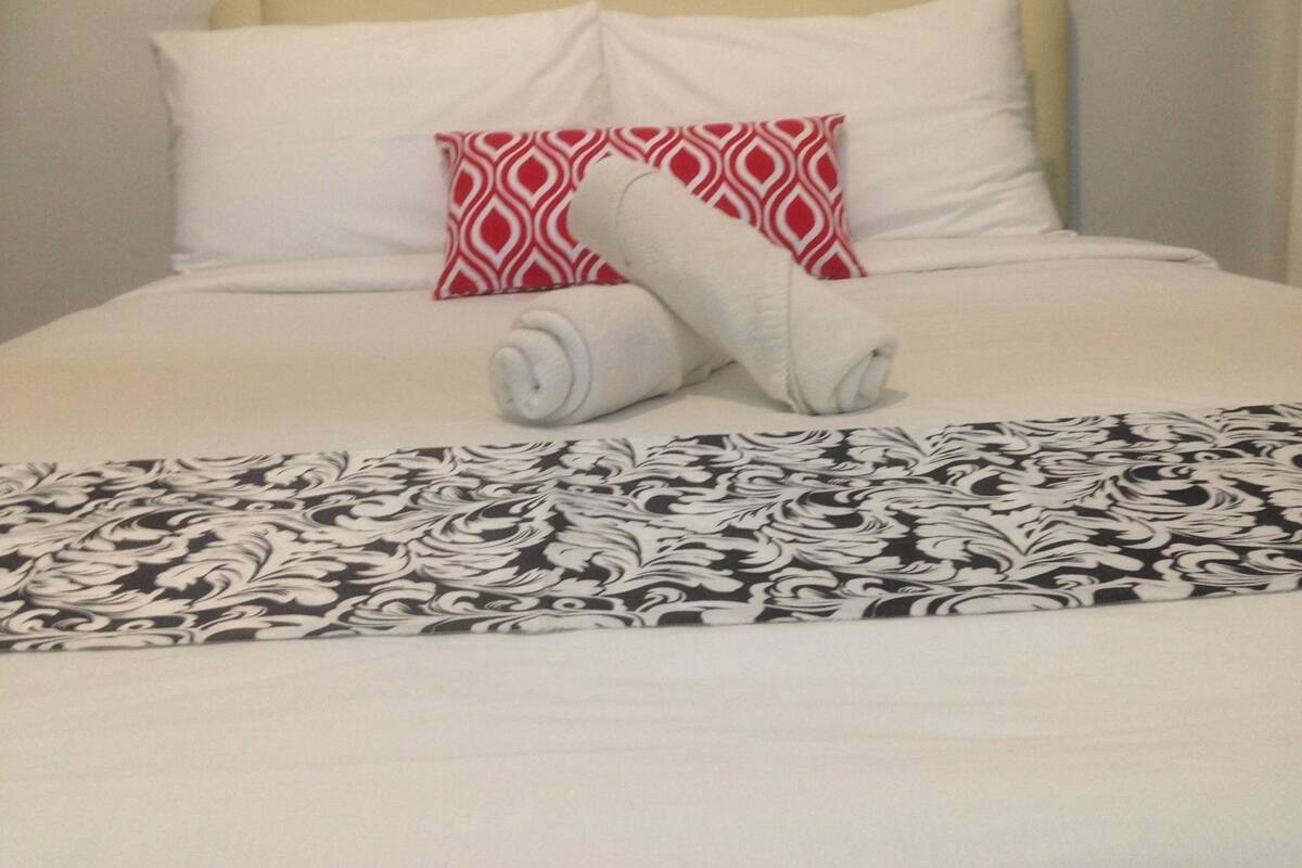 A bed with white bedding, a red and white pillow, and a rolled white towel.
