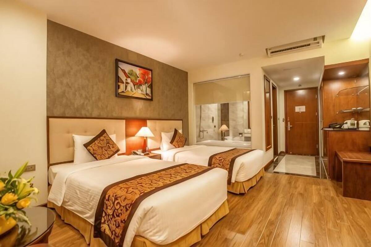 A hotel room with two beds, a wooden floor, a painting on the wall, a mirror, and a bathroom door.