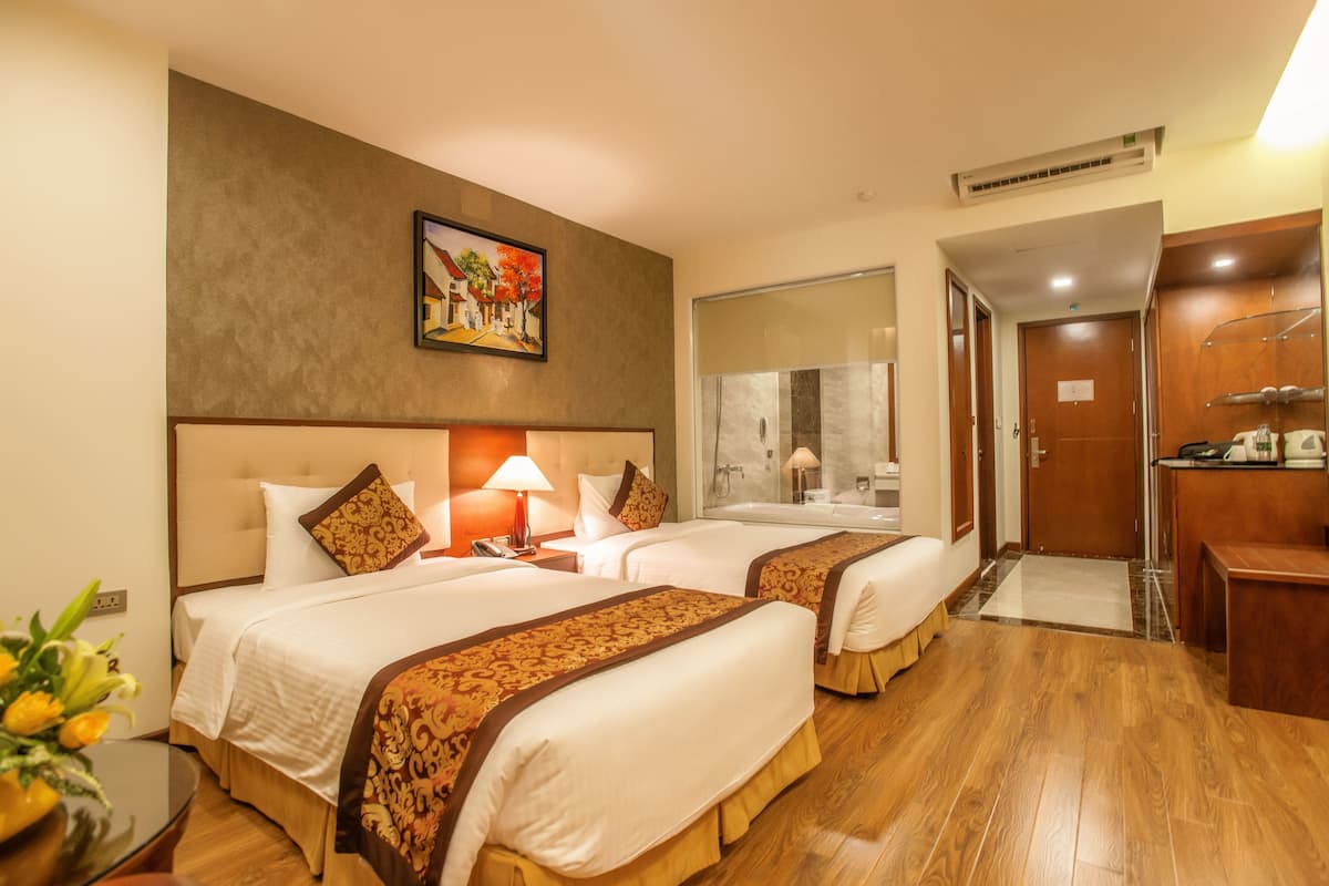 A hotel room with two beds, a painting on the wall, a bathroom with a glass door, and a desk with a chair.