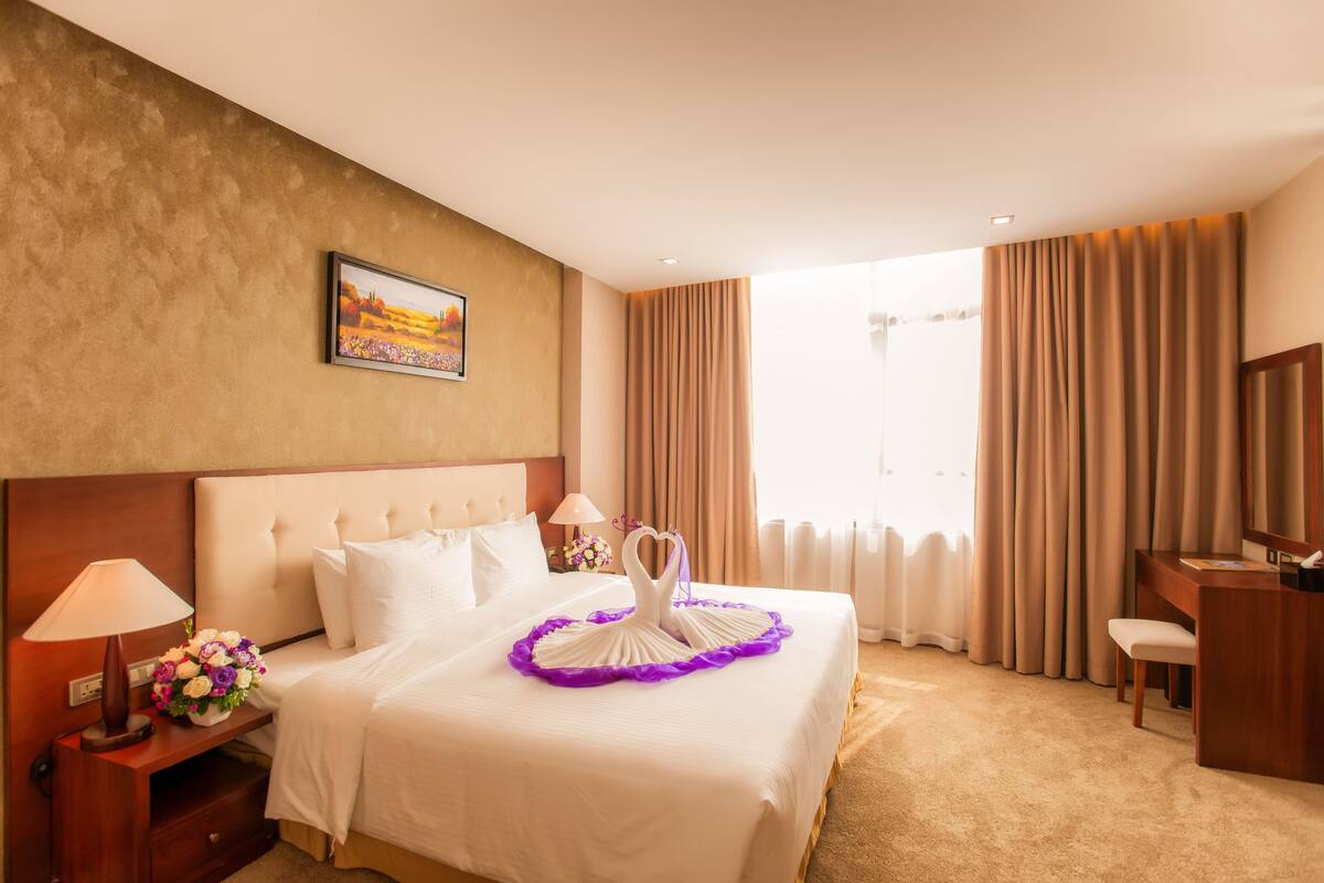A hotel room with a large bed, a desk, a chair, a nightstand with a lamp, a bouquet of flowers, and a painting on the wall.