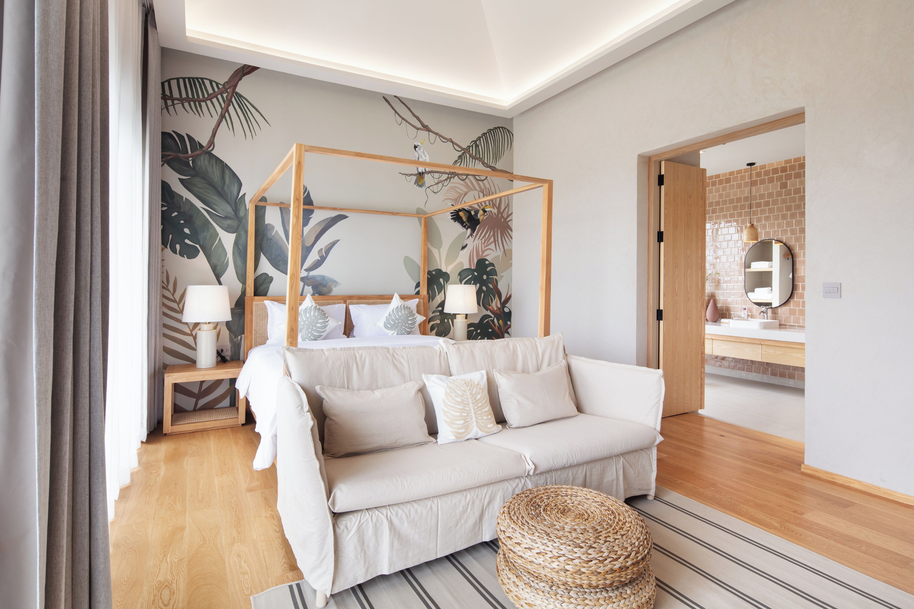 A modern bedroom with a white sofa, a wooden canopy bed, a large mirror, and a tropical leaf mural.
