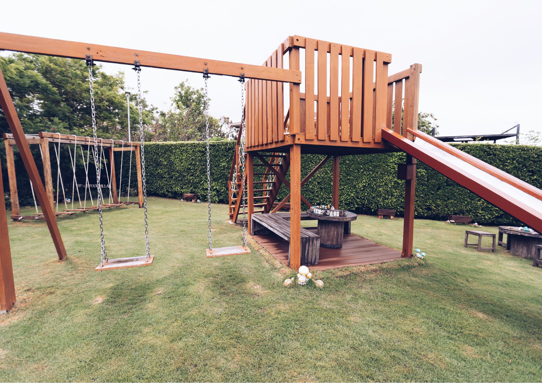 A wooden playground structure with swings and a slide, surrounded by a well-maintained lawn and a hedge.