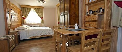 Un dormitorio con piso de madera, una cama, una mesa de comedor de madera con sillas, un armario con utensilios de cocina y una ventana con cortinas.