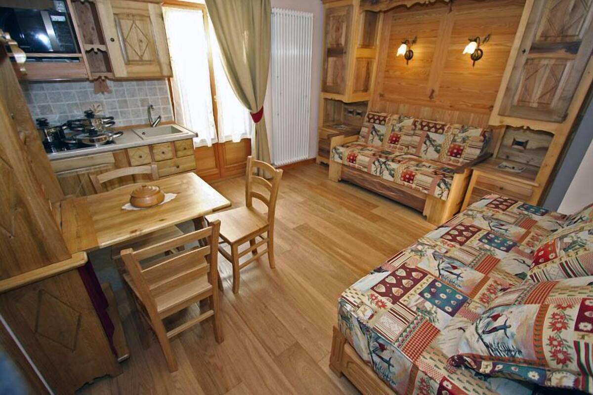 A cozy kitchen with wooden cabinets, a dining table, and chairs. There is a sofa with a quilted cover and a window with curtains.
