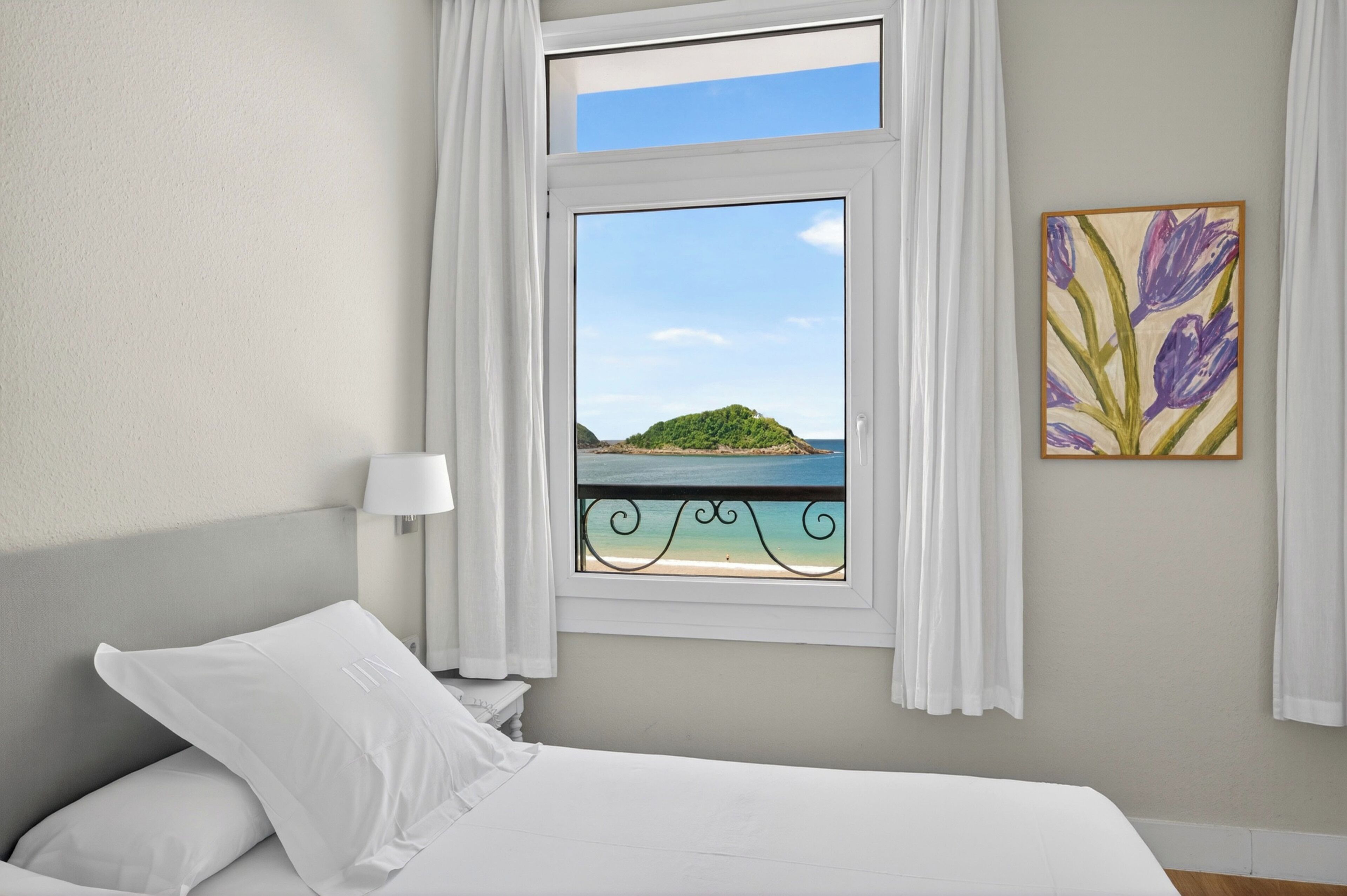 Un dormitorio con cama, ventana que ofrece vistas a una playa y una isla, mesita de noche con lámpara y un cuadro colgado en la pared.
