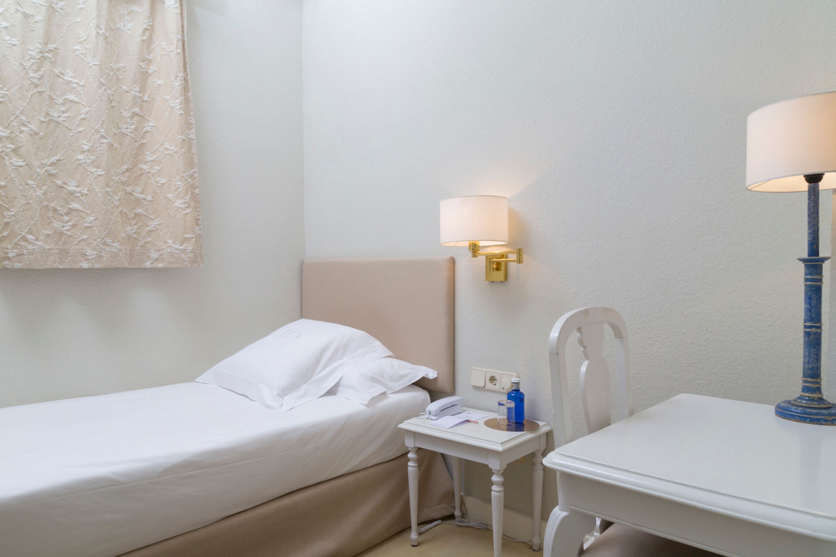 A hotel room with a single bed, a bedside table with a lamp, a chair, and a small table with a phone and a bottle.