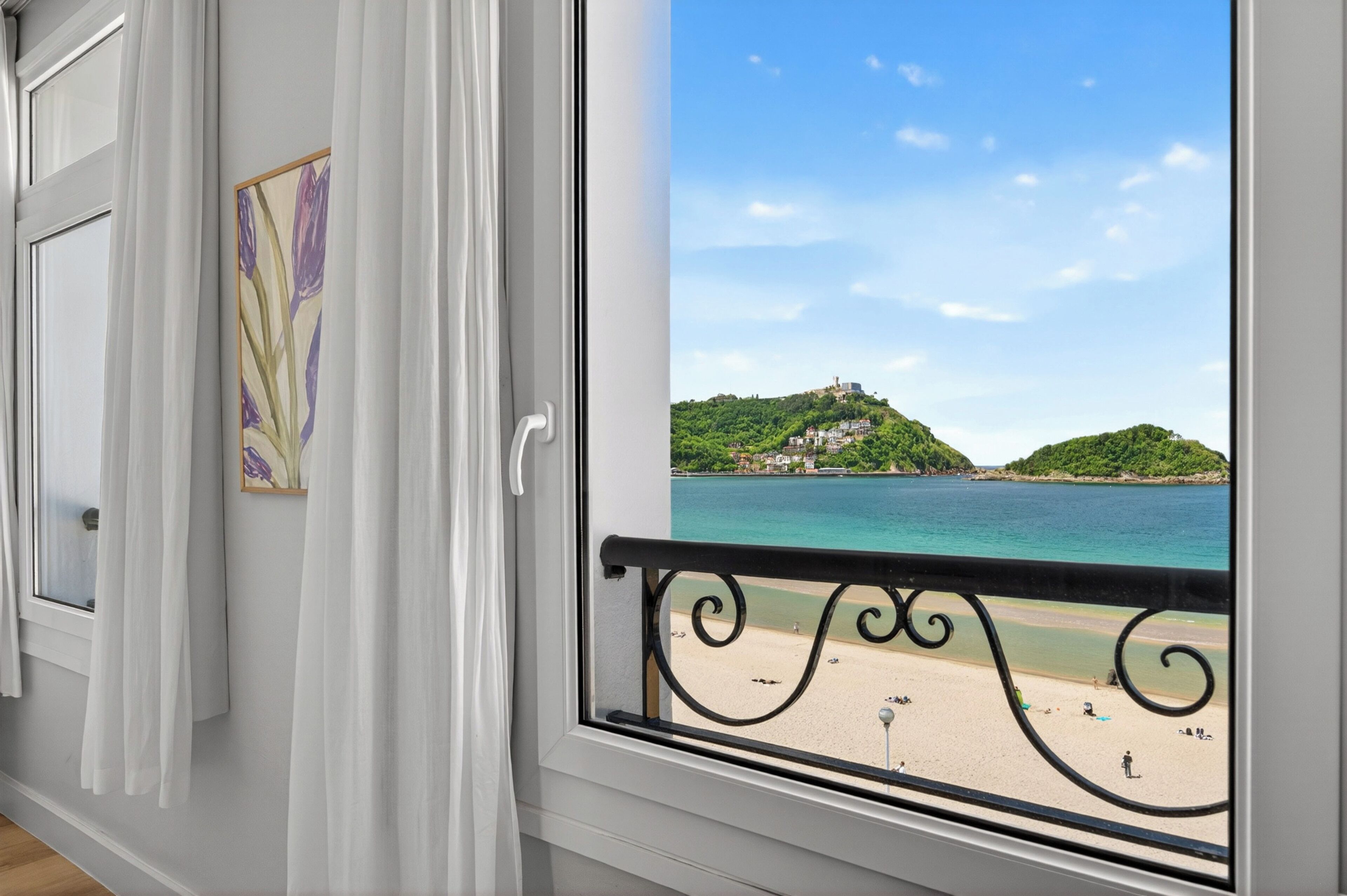 A room with a large window overlooking a beach, a balcony with a metal railing, and a framed picture on the wall.