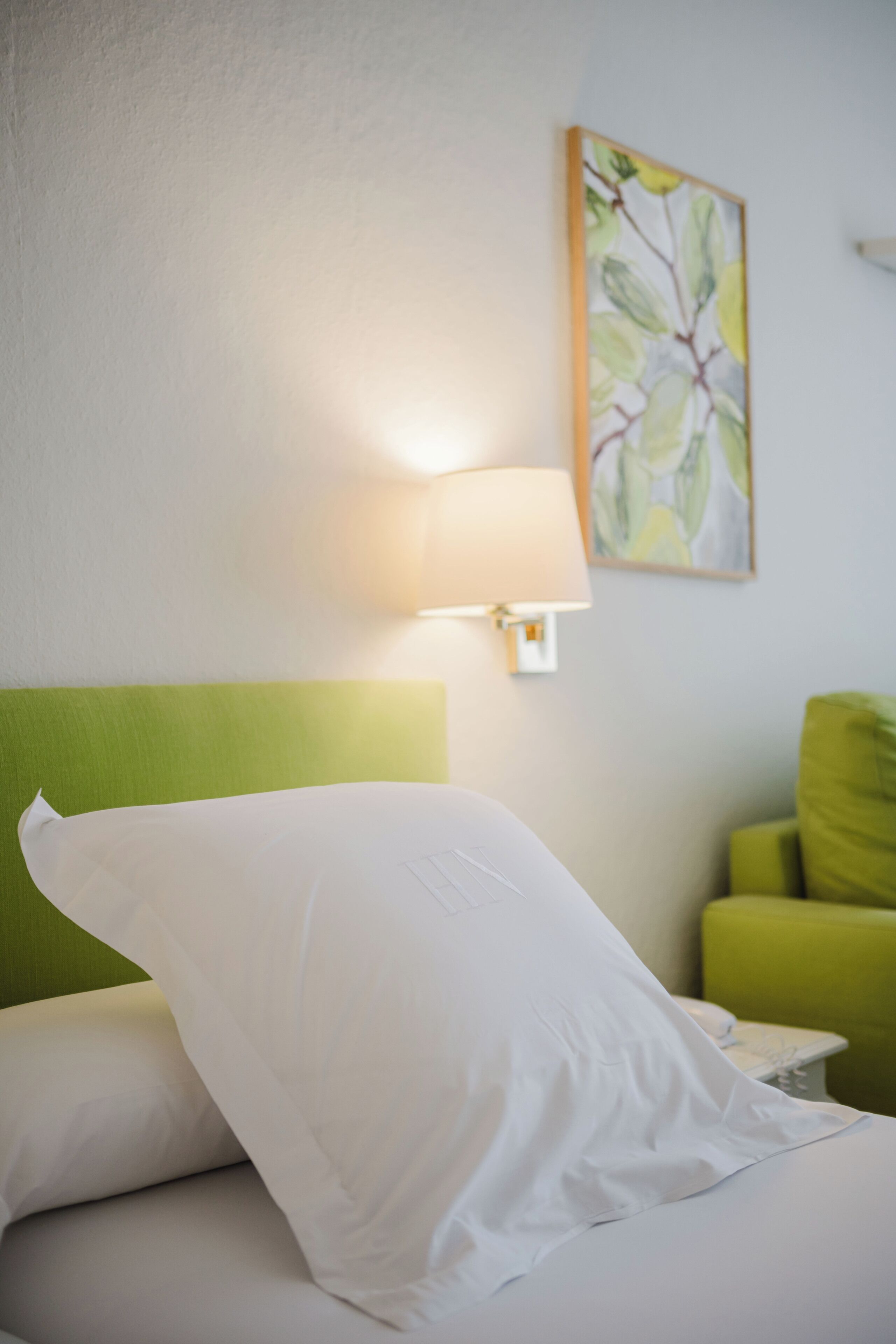 Una cama bien hecha con sábanas blancas, un cabecero verde, una lámpara fijada a la pared y un sillón verde.