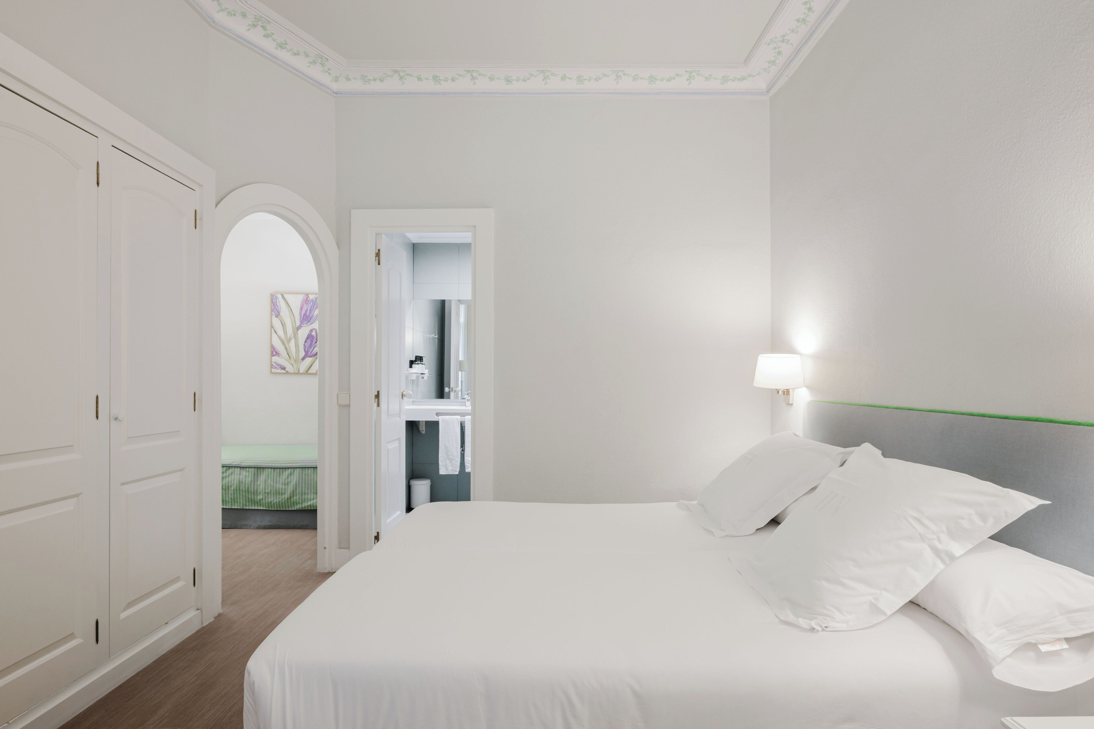 A hotel room with a bed, a nightstand, a lamp, a wardrobe, and an open door leading to a bathroom.