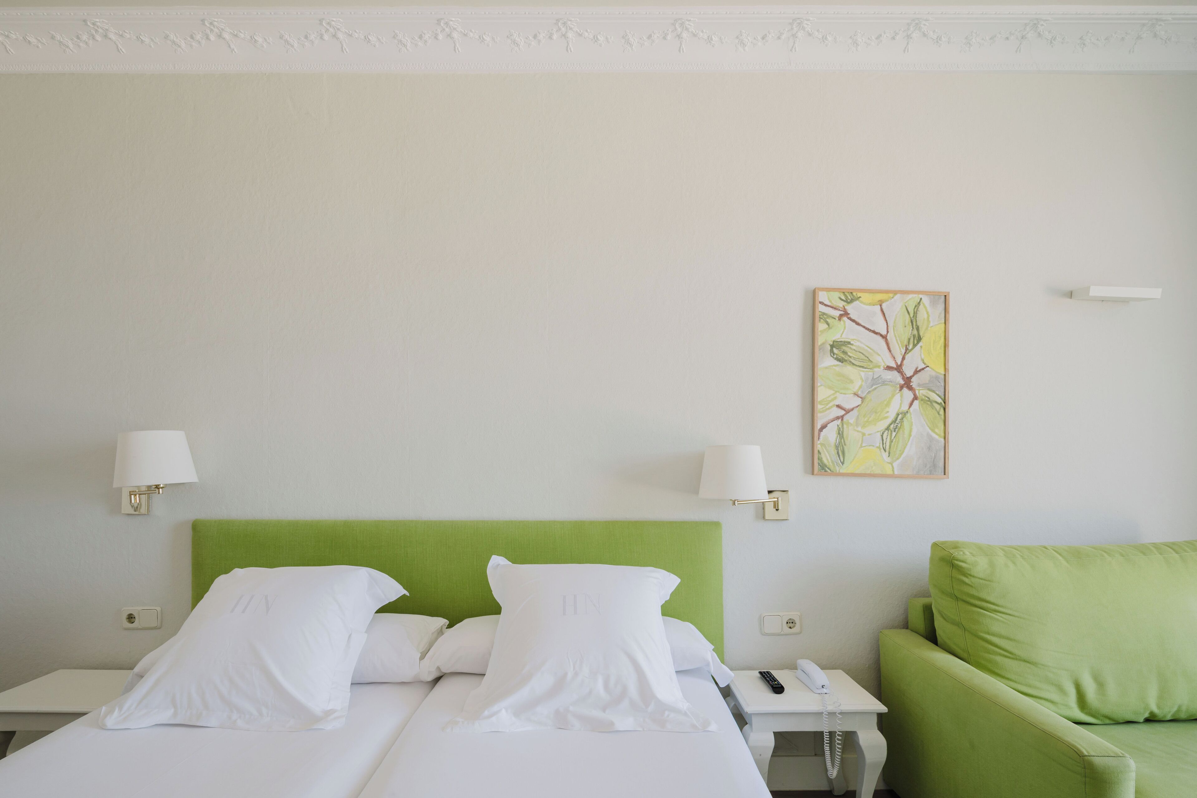 A hotel room with two beds, a green headboard, white bedding, a green sofa, a painting on the wall, and a lamp on each side of the beds.
