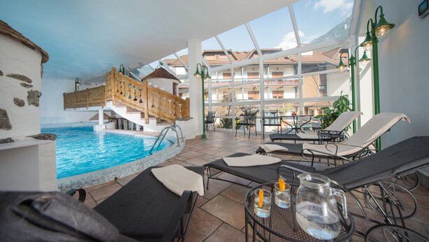 An indoor swimming pool area with lounge chairs and a wooden staircase leading to upper floors. - Hotel Mühlener Hof (Campo Tures)
