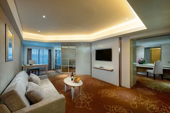 A modern hotel room with a sofa, a small table with a fruit bowl, a TV, and a desk with a chair. - Rio Hotel & Casino (Macau)