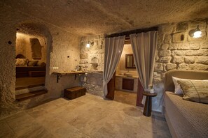 Un interior rústico de piedra con una cama, un banco de madera, una mesita y una pared de piedra con estante empotrado.