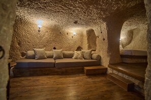 A cozy interior with a stone wall, a wooden bench, and a sofa with cushions. - Meleklerevi Cave Hotel (Ürgüp)